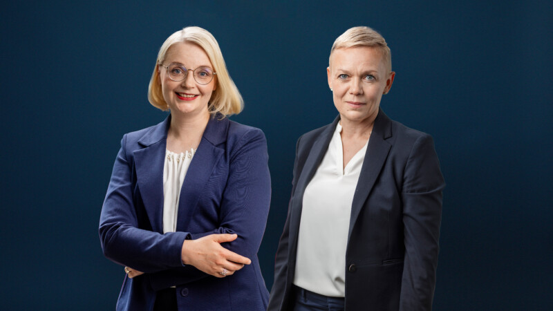 Kaksi naista tummansinistä taustaa vasten; vasemmanpuoleinen vaaleahiuksinen hymyilee silmälasit päässä ja pitää käsiään edessään, oikeanpuoleinen lyhyttukkainen katsoo vakavana.