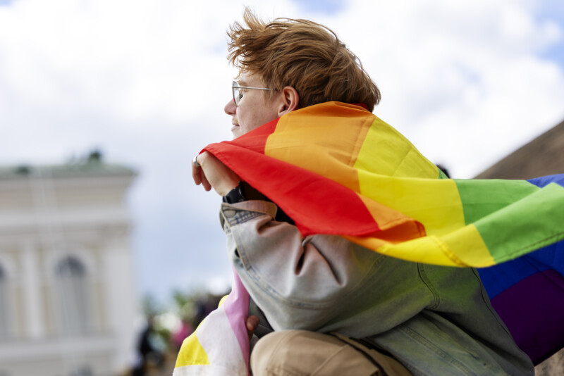 Nuori henkilö sivuprofiilissa katsomassa kauas, hartioilla suuri sateenkaarilippu Pride-teemassa, taustalla kaupungin rakennuksia ja pilvinen taivas.