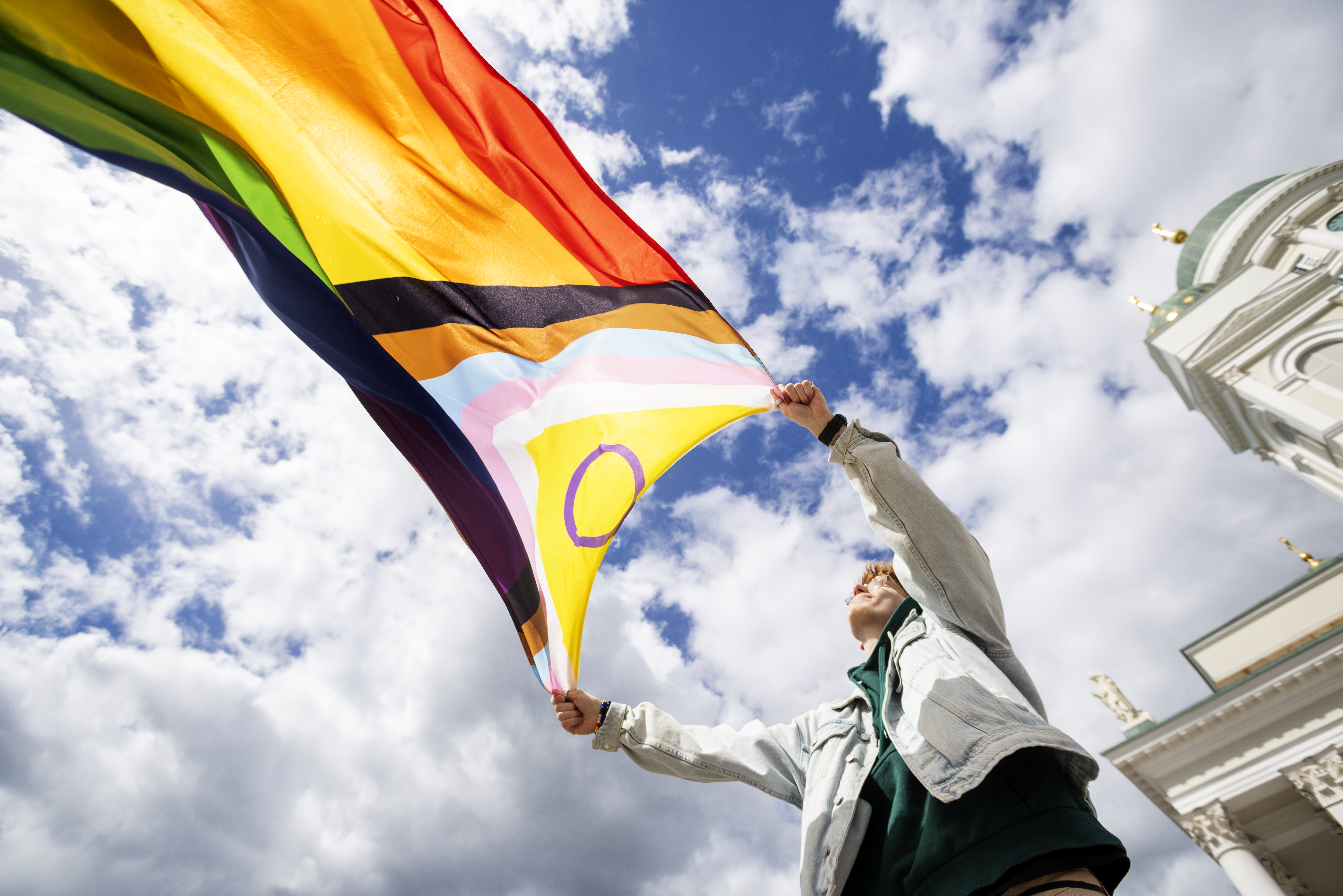Alaviistosta otettu kuva, jossa henkilö pitää suurta Progress Pride -lippua liehumassa sinistä taivasta ja pilviä vasten Helsingin tuomiokirkon edessä.