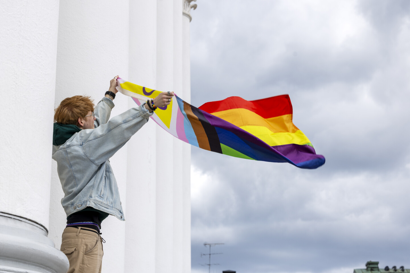 Henkilö seisoo valkoisen rakennuksen pylvään reunalla ja pitää käsissään tuulessa liehuvaa Progress Pride -lippua, jossa näkyvät sateenkaari-, trans- sekä mustat ja ruskeatraidat, harmaa pilvinen taivas taustalla.