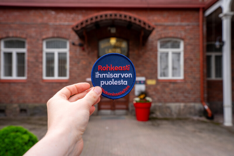 Etualalla käsi pitää sinistä pyöreää rintanappia, jossa valkoisella ja punaisella lukee "Rohkeasti ihmisarvon puolesta", taustalla punatiilinen rakennuksen sisäänkäynti ja kukkapotti.
