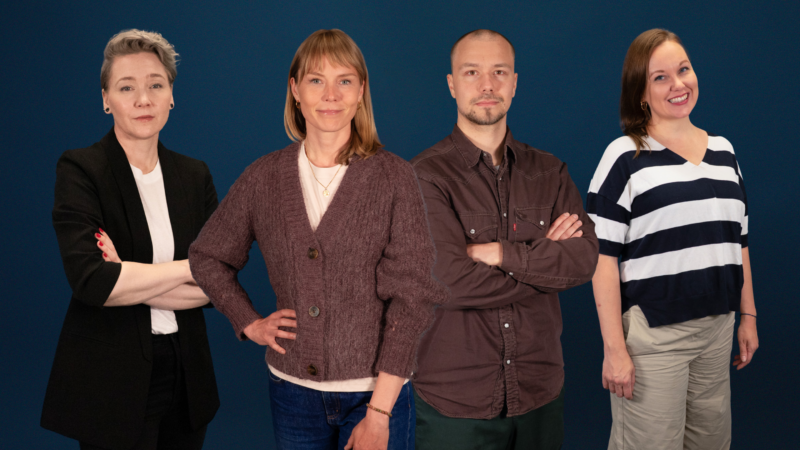 Neljä aikuista seisoo vierekkäin tummansinisen taustan edessä; heillä on rento‑ammattimainen vaatetus ja ystävälliset, itsevarmat ilmeet.