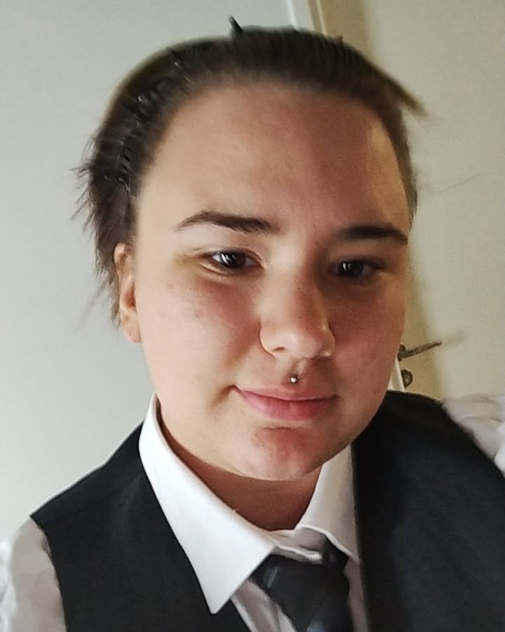 Lähikuva henkilöstä, jolla on tummat hiukset ponnarilla, valkoinen kauluspaita, tumma liivi ja solmio, nenänlävistys ja rauhallinen ilme.