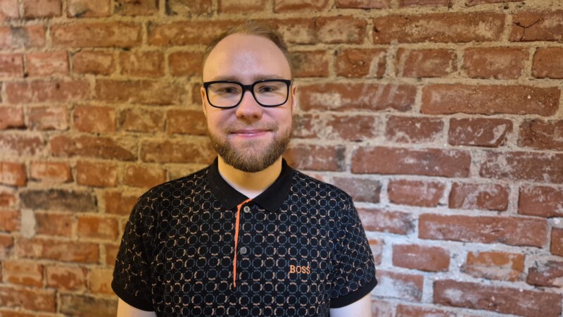 Henkilö seisoo tiiliseinän edessä ja katsoo kameraan