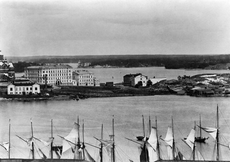 Mustavalkoinen historiallinen panoraamakuva: etualalla merivesi ja rivissä purjelaivoja mastoineen, taustalla Katajanokan ranta-alue vanhoine kivirakennuksineen ja kaupunkimaisemineen.