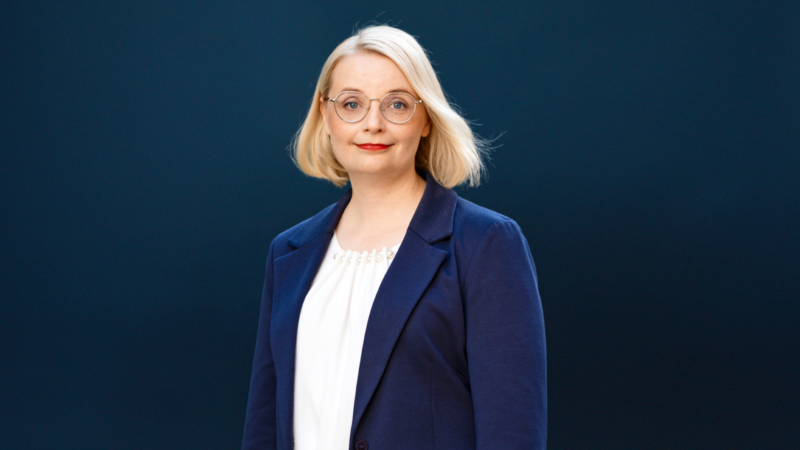 Vartalonmittainen muotokuva vaaleahiuksisesta naisesta, joka katsoo kameraan ja hymyilee, käyttää pyöreitä laseja, valkoista paitaa ja sinistä bleiseriä, taustalla tasainen tumma väri.