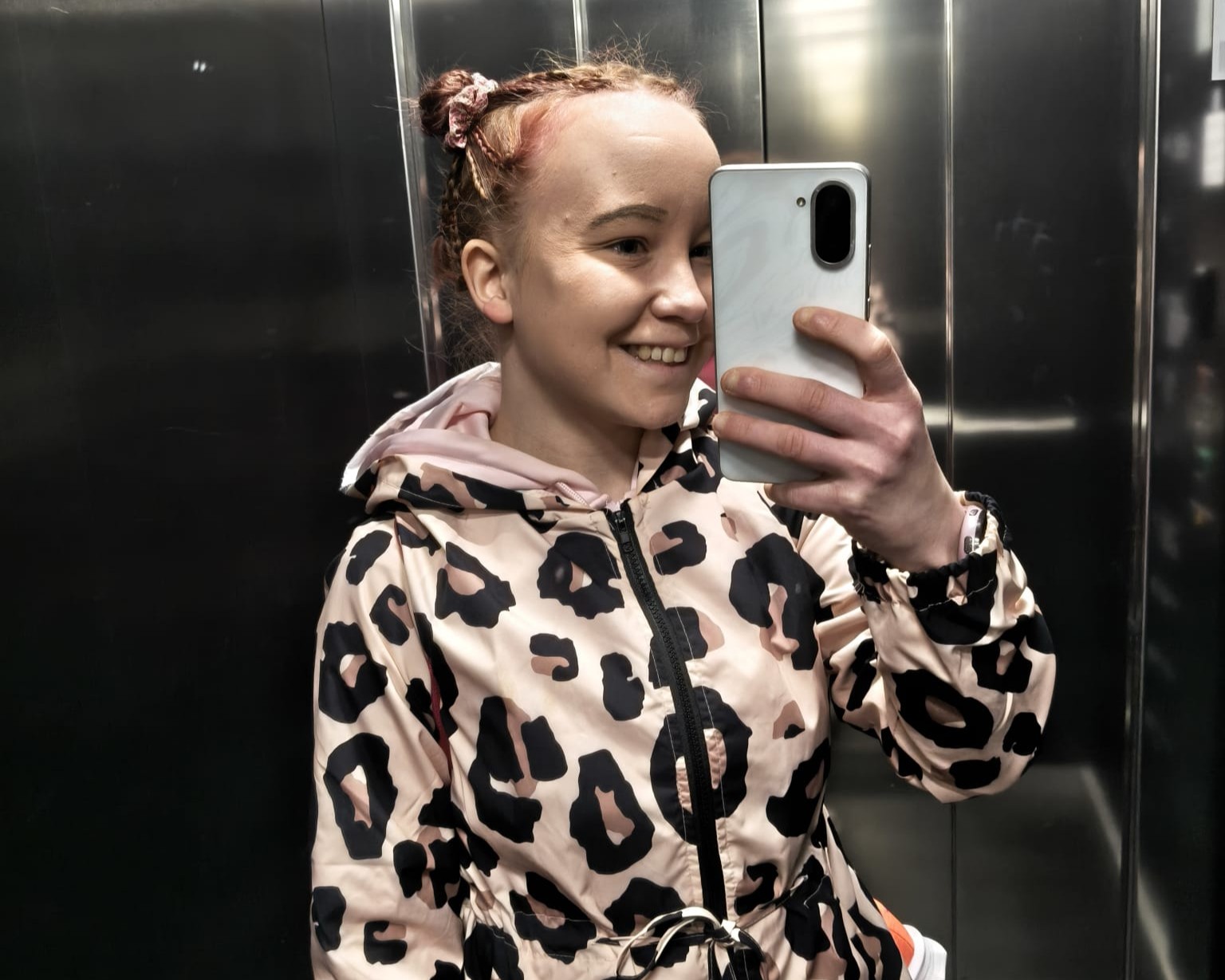 Henkilö ottaa hississä peilistä selfien ja hymyilee, pukeutunut vaaleanpunaiseen leopardikuvioiseen huppariin, hiukset nutturoilla ja pitää valkoista älypuhelinta, taustalla metalliset hissiseinät.