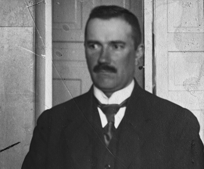 Black-and-white early 20th-century portrait of a middle-aged man with dark hair and a trimmed mustache, wearing a dark suit, vest and tie, standing indoors against a plain paneled wall and looking slightly to the right with a serious expression.