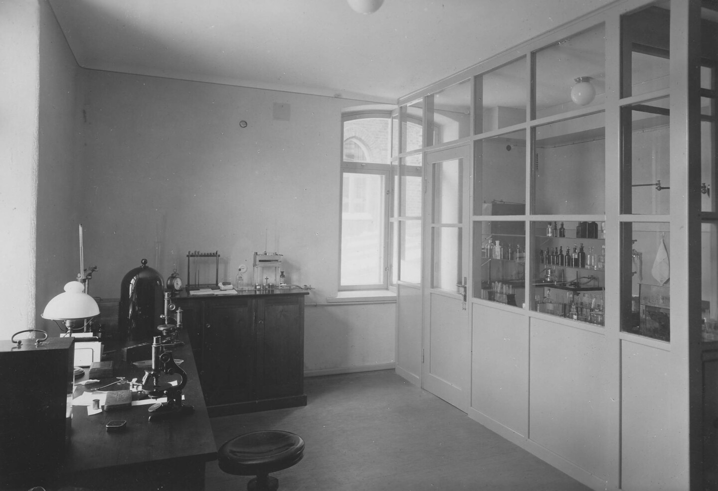 Black-and-white interior photograph of an empty hospital laboratory showing a workbench with a microscope and lamp, stools and equipment, cabinets and glass bottles visible through a glass-partitioned room, and a tall window letting in daylight.
