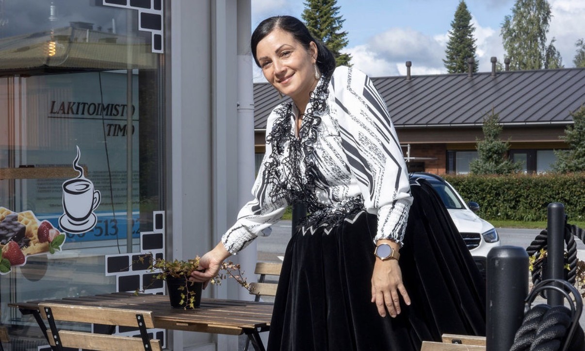 Hymyilevä nainen perinteisessä romaniasussa asettaa kukkaruukkua kahvilan ulkopöydälle terassilla, taustalla kahvilan ikkuna, taloja, auto ja puita.