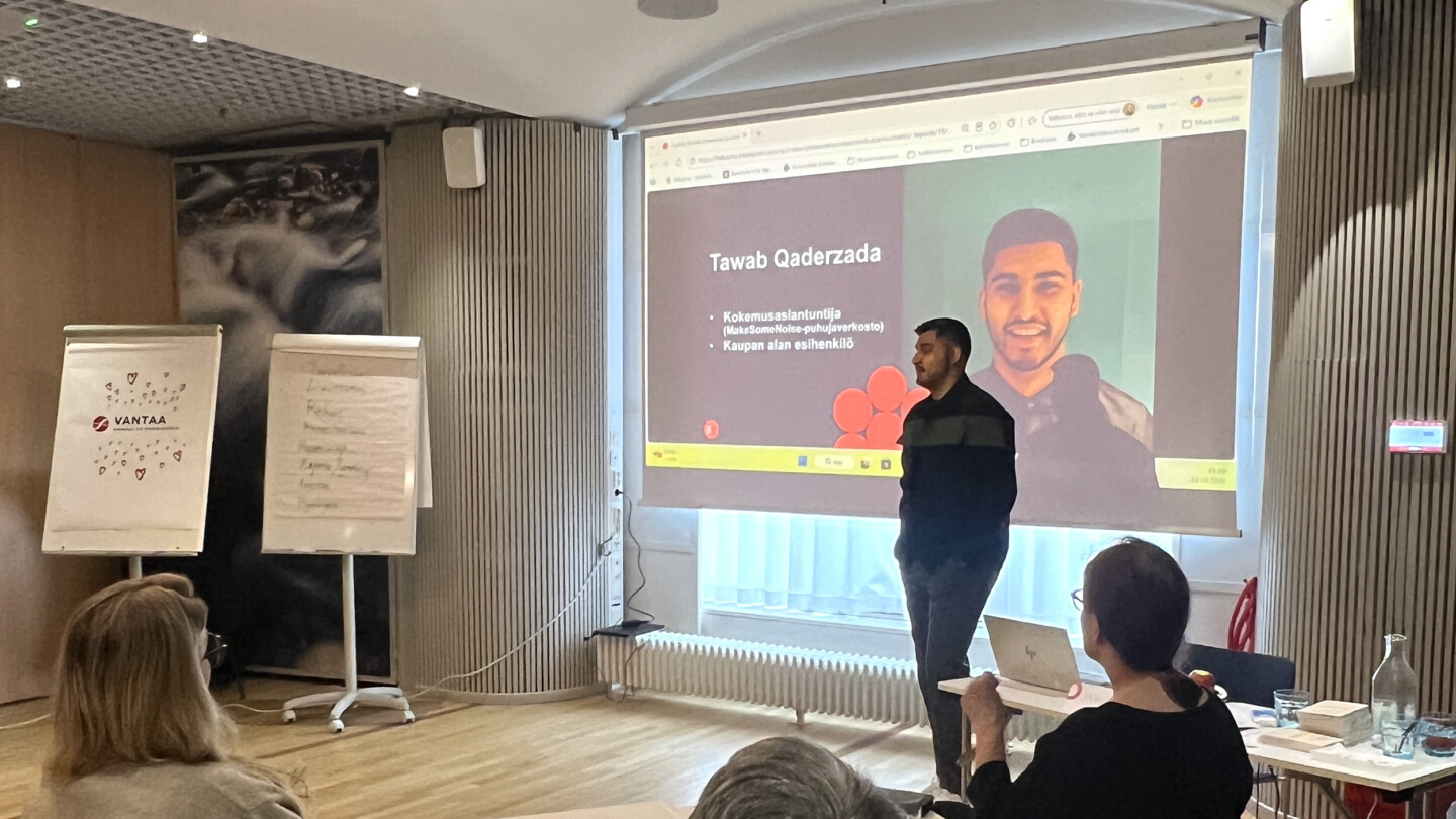 Esittelijä seisoo kokoushuoneessa projektorin edessä näyttäen diaa; muut osallistujat istuvat katsomassa ja vasemmalla näkyy kaksi fläppitaulua.
