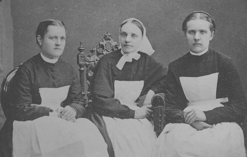 Mustavalkoinen historiallinen valokuva kolmesta diakonissapuvuissa olevasta naisesta istumassa vierekkäin; heillä on tummat puvut, valkoiset esiliinat ja päähineet, taustalla koristeltu tuoli.